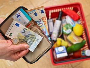 Eine Frau hält auf ihrer Hand Geld vor einem vollen Einkaufskorb mit Lebensmitteln.  
