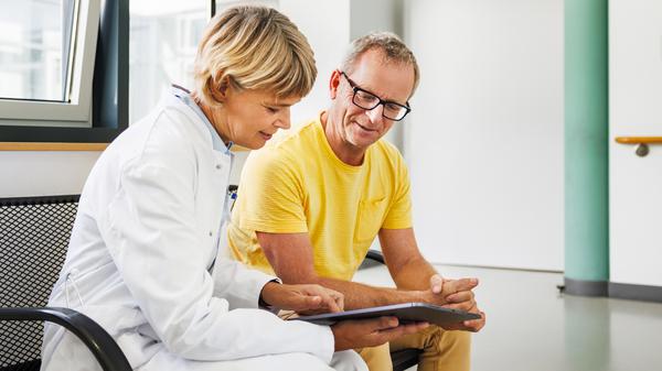 Ein mittelalter Mann sitzt auf einer Bank neben einer Ärztin, die ihm etwas auf einem Tablet zeigt.