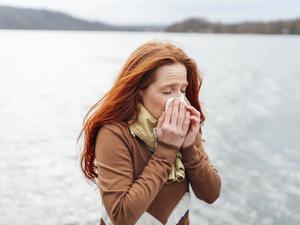 Eine erkältete Frau schnäuzt sich die Nase, hinter ihr Seepanorama.