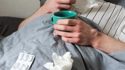 Ein Mann liegt mit Taschentüchern, Teetasse, Nasenspray und Tabletten im Bett (gestellte Szene).