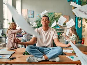 Ein junger Mann meditiert sitzend auf einem Schreibtisch, während um ihn im Büro Chaos herrscht.