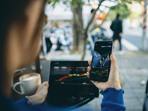 Eine Frau sitzt in einem Café und schaut auf Smartphone und Laptop Aktienkurse an.