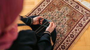 Beautiful young Muslim woman praying using prayer beads (misbaha,tasbih) and a prayer mat at home. Close-up.