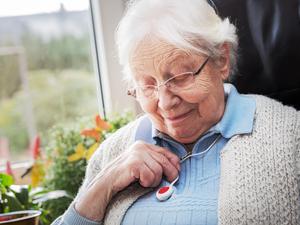 Eine ältere Dame blickt auf den Notrufknopf, den sie an einer Kette um den Hals trägt.