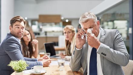 Ein Mann im Anzug niest in ein Taschentuch, während seine Kollegen im Hintergrund ihn erschrocken anschauen (Symbolfoto).