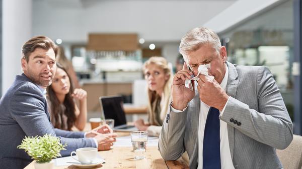 Ein Mann im Anzug niest in ein Taschentuch, während seine Kollegen im Hintergrund ihn erschrocken anschauen (Symbolfoto).