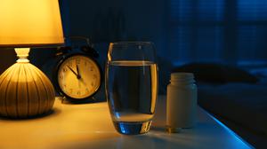 Illustration: Eine Tablettenbox, ein Glas Wasser und ein Wecker stehen auf einem Nachttisch.
