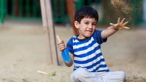 Ein etwa fünfjähriger Junge sitzt auf einem Spielplatz und wirft mit Sand. Er trägt ein blau gestreiftes T-Shirt.