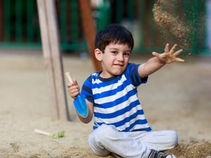 Ein etwa fünfjähriger Junge sitzt auf einem Spielplatz und wirft mit Sand. Er trägt ein blau gestreiftes T-Shirt.