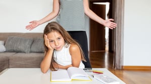 Ein etwa zehnjähriges Mädchen sitzt gestresst vor ihren Unterlagen am Wohnzimmertisch. Hinter ihr breitet ihre Mutter resigniert die Arme aus.