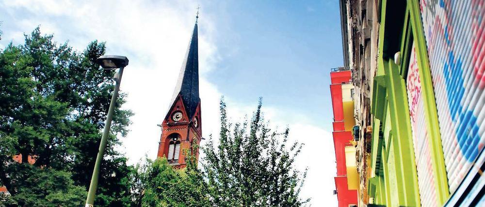 Immanuelkirchstraße in Prenzlauer Berg: Einfallstor und Trutzburg. Blick auf die Immanuelkirche und den oberen Abschnitt der Immanuelkirchstraße (Richtung Prenzlauer Allee). Rechts erkennt man die roten Erker des Immanuelkirch-Carrés. Dieser 2008 fertiggestellte Komplex ist Inbegriff für die Gentrifizierung der Straße.
