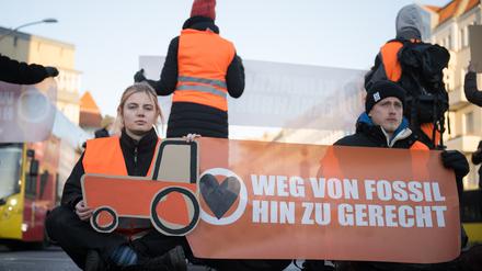Aktivisten der Klimaschutzgruppe Letzte Generation sitzen bei einer Straßenblockade auf dem Innsbrucker Platz mit einem gemalten Traktor und einem Banner „Weg von fossil hin zu gerecht“ auf der Straße.