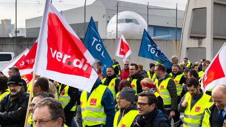 Ein Protestzug von Streikenden zieht mit Bannern und Verdi-Fahnen vom Lufthansa Aviation Center zum Terminal 1. Im Hintergrund ist ein Flugzeug der Lufthansa zu sehen.
