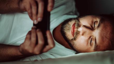 Ein junger Mann liegt im Bett und schaut auf ein Smartphone.