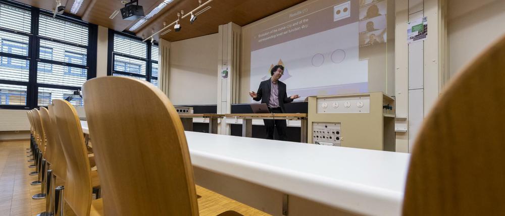 Ein Professor steht in einem leeren Hörsaal vor einer Leinwand, auf der Vorlesungsinhalte und weitere Teilnehmende zu sehen sind.