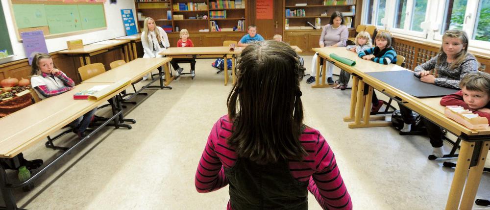 Eine Schülerin im Grundschulalter steht in einem Klassenraum vor neun Mitschülern verschiedenen Alters und zwei Lehrerinnen.