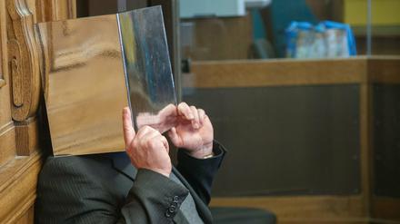 Der Angeklagte sitzt auf der Anklagebank und hält sich einen Aktenordner vor das Gesicht. 