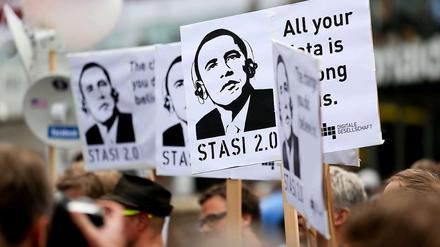 Demonstranten halten bei Protesten während des Besuchs von Barack Obama in Berlin am Checkpoint Charlie Plakate hoch, auf denen Obama mit Kopfhörern zu sehen ist.