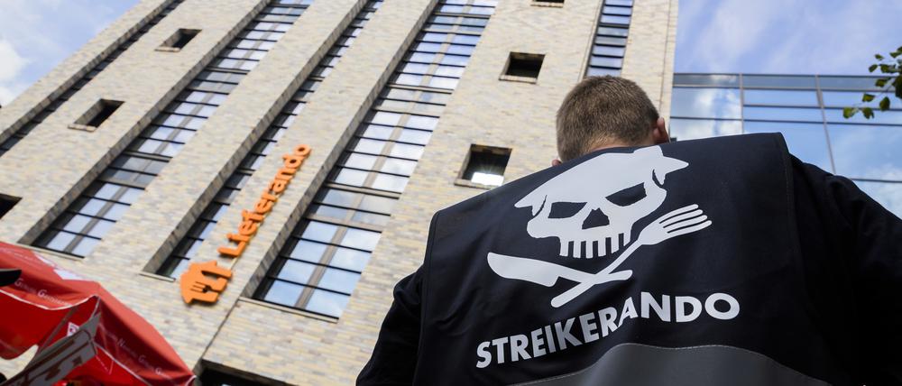 17.08.2023, Berlin: Ein Teilnehmer steht bei einer Kundgebung zum Streik von Beschäftigten des Essenslieferdienstes Lieferando für bessere Bezahlung vor dem Firmensitz von Lieferando. Dabei trägt er eine Veste mit einem zum Totenkopf umgestalteten Lieferando-Logo. Die Gewerkschaft Nahrung-Genuss-Gaststätten (NGG) hat Lieferando-Beschäftigte aus ganz Deutschland zum Streik aufgerufen. Foto: Christoph Soeder/dpa +++ dpa-Bildfunk +++