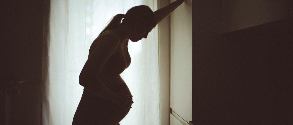 Eine schwangere Frau, die eher als Silhouette vor einem Fenster zu sehen ist, stützt sich mit der Hand an einem Schrank ab, um eine Wehe besser zu ertragen (Symbolbild).