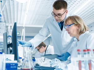 In einem modernen Labor führt eine erfahrene Wissenschaftlerin ein Gespräch mit einem jungen Laborassistenten. Er zeigt ihr Diagramme auf einem Klemmbrett, sie analysiert die Daten und gibt sie in ihren Computer ein.
