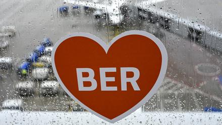 Ein Aufkleber mit einem Herz und den Buchstaben BER ist auf der Besucherterrasse am Flughafen BER bei Regenwetter vor einem Flugzeug zu sehen.