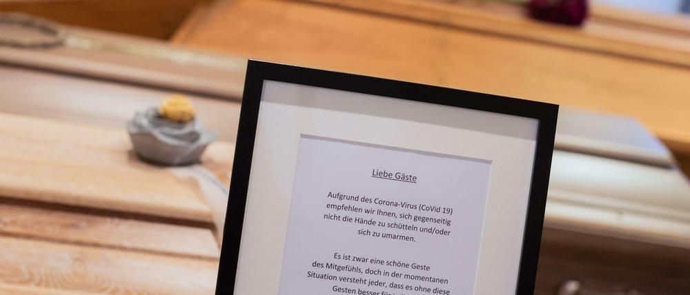 Ein Hinweisschild für den Umgang bei Beerdigungen steht in einem Bestattungsinstitut vor mehreren Särgen. Der Hinweis informiert, dass Trauernde sich aufgrund des Coronavirus' nicht die Hände zu schütteln oder umarmen sollen.