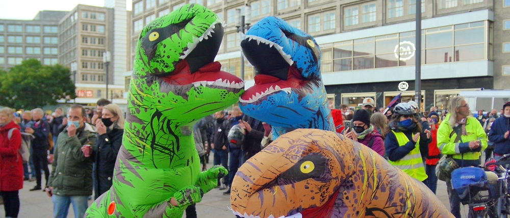 Die Echsenmenschen stören Verschwörungsgläubige auf dem Alexanderplatz. Beni steckt im grünen Kostüm.