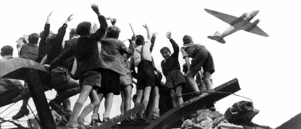 West-Berliner Jungen, die auf einem Trümmerberg stehen, begrüßen winkend ein US-amerikanisches Transportflugzeug.