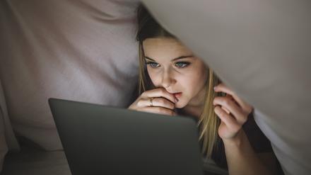 Eine junge Frau schaut unter der Bettdecke liegend auf den Bildschirm ihres Laptops.
