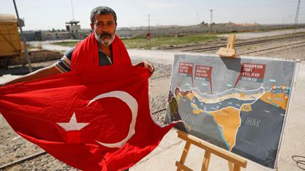 Ein Mann mit einer türkischen Flagge steht an der Grenze zwischen der Türkei und Syrien neben einer Karte, welche die geplante militärische Operation der Türkei in Syrien zeigt.