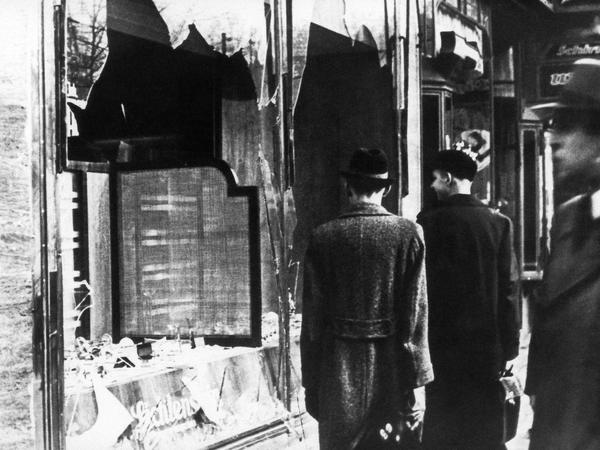 Passanten gehen an einem Schaufenster mit eingeschlagenen Scheiben vorbei. Ein Schwarz-Weiß-Foto aus den 1930er Jahren.