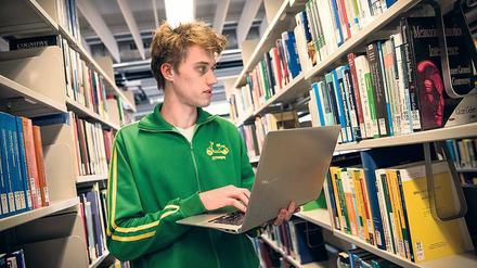 Ein Student steht mit aufgeklapptem Laptop zwischen den Regalen einer Bibliothek.