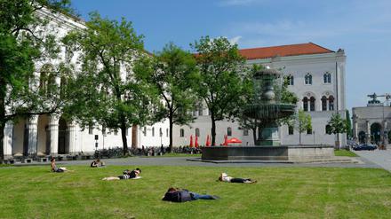 Vor dem historischen Hauptgebäude der LMU München liegen Studierende auf dem Rasen und lesen.